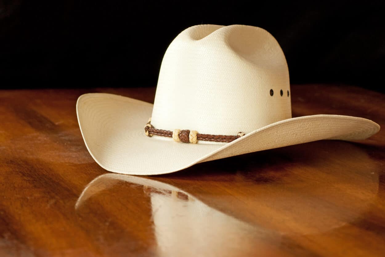 White straw cowboy hat with a hatband on a wooden table against dark background