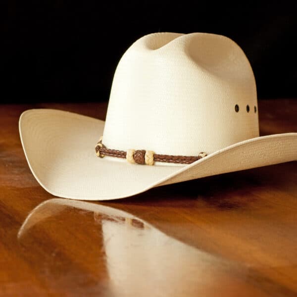 White straw cowboy hat with a hatband on a wooden table against dark background